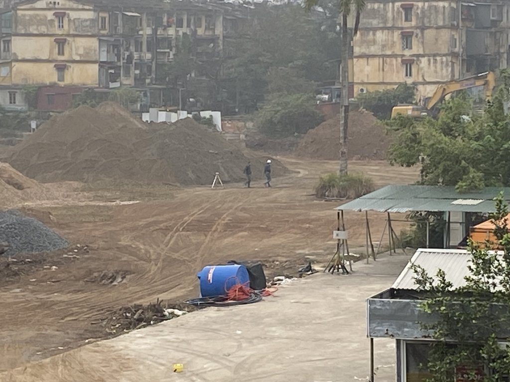 Tiến Độ Xây Dựng Vinhomes Giảng Võ - Tháng 4/2025 9 Một khu đất rộng tại dự án Vinhomes Giảng Võ đã được san lấp, với các máy xúc và vật liệu xây dựng, phía xa là các tòa nhà cao tầng và một con đường chạy dọc công trường.