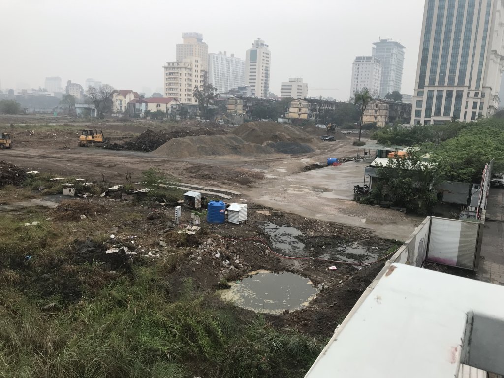 Tiến Độ Xây Dựng Vinhomes Giảng Võ - Tháng 4/2025 8 Công trường dự án Vinhomes Giảng Võ với máy xúc đang làm việc, các đống đất lớn và vật liệu xây dựng, xung quanh là các tòa nhà cũ và cao tầng trong không khí mờ sương.