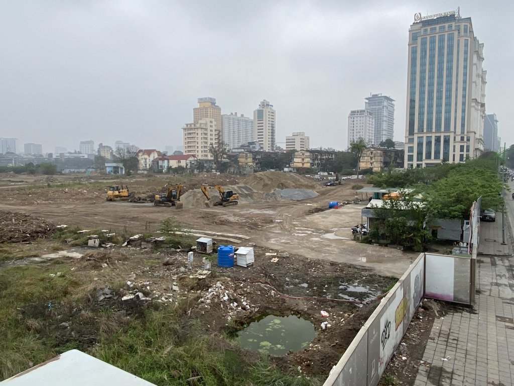 Tiến Độ Xây Dựng Vinhomes Giảng Võ - Tháng 4/2025 5 Công trường dự án Vinhomes Giảng Võ với các máy xúc và máy ủi đang làm việc, các đống đất và vật liệu xây dựng được tập kết, xung quanh là các tòa nhà cao tầng trong không khí mờ sương.