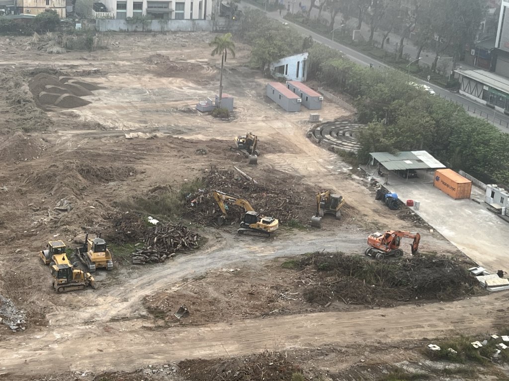Giá bán Vinhomes Giảng Võ - Bảng giá và chính sách mới nhất 2025 5 Một khu đất tại dự án Vinhomes Giảng Võ với các máy xúc, đống đất và vật liệu xây dựng, bên cạnh là các tòa nhà cao tầng và một con đường chạy dọc công trường, trong không khí mờ sương.