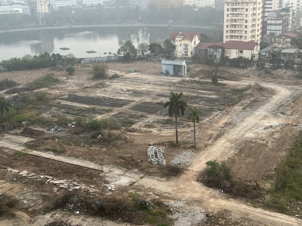 Tiến Độ Xây Dựng Vinhomes Giảng Võ - Tháng 4/2025 3 Công trường thi công dự án Vinhomes Giảng Võ với máy xúc đang làm việc, các đống đất lớn và vật liệu xây dựng, phía xa là các tòa nhà cao tầng và cây cọ, trong không khí mờ sương.